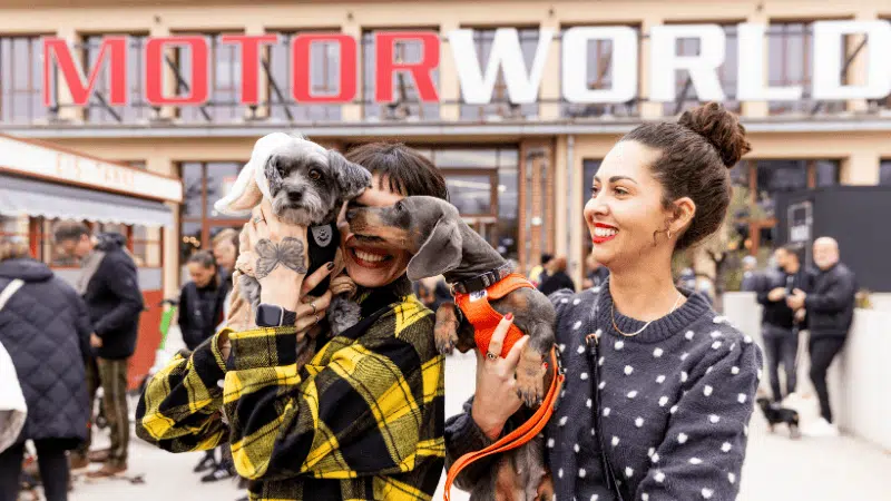 2 Frauen halten jeweils einen Dackel und einen kleinen, grauen Mischlingshund im Arm. Im Hintergrund sieht man den Eingangsbereich der Motorworld in München.